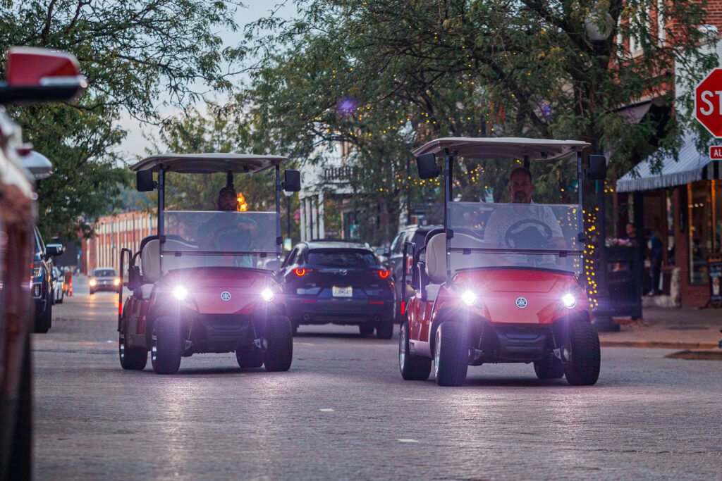 Two friends driving red and orange golf carts for rent are experiencing Historic Main Street nightlife